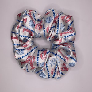 Op de afbeelding: Een witte satijnen scrunchie met een patroon van roze kreeften, blauwe zeeschelpen en blauwe golvende lijnen. Een haaraccessoire.