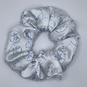 Puede incluir: Una goma para el pelo de satén blanco con un patrón repetido de diseños de dólares de arena azules. La tela está fruncida, creando una apariencia voluminosa y texturizada. La goma es un accesorio para el cabello.