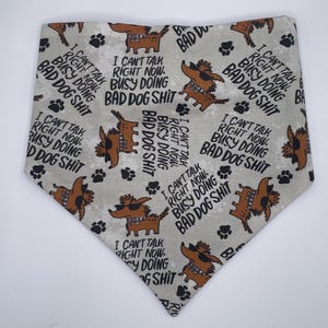 May include: A triangular pet bandana in light gray featuring a repeating pattern of brown cartoon dogs wearing sunglasses and collars, interspersed with black paw prints and the text I CAN'T TALK RIGHT NOW. BUSY DOING BAD DOG SHIT.