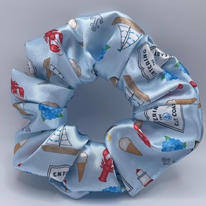 Op de afbeelding: Een lichtblauwe stoffen haarelastiek met een herhalend patroon van rode kreeften, zeilboten, ijshoorntjes, blauwe hortensia's, ankers en bordjes met de tekst Entering the East Coast met een klein ankericoontje in het midden.