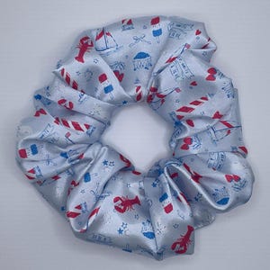 Op de afbeelding: Een lichtblauwe stoffen haarelastiek met een herhalend patroon van rode, witte en blauwe illustraties, waaronder kreeften, zeilboten, ijsjes, kersen en banners met de tekst RED, WHITE, AND BLUE verspreid over het oppervlak.