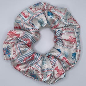 Puede incluir: Una scrunchie de satén blanco con un estampado repetido de langostas rojas y azules, corales y diseños geométricos. La scrunchie tiene forma circular, perfecta para asegurar el cabello en una cola de caballo o moño.