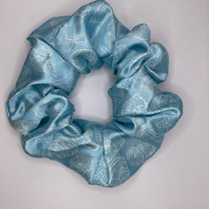 Puede incluir: Una goma para el pelo de satén azul claro con un estampado de conchas blancas. El accesorio para el cabello es redondo y fruncido, con una textura suave y brillante. La goma es un accesorio para el cabello.