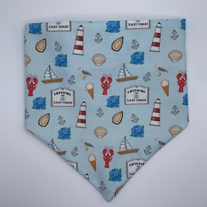 Op de afbeelding: Een lichtblauwe, driehoekige stoffen bandana met een herhalend patroon van rood-witte vuurtorens, zeilboten, rode kreeften, blauwe hortensia's, schelpen, meeuwen, ijshoorntjes, ankers en bordjes met de tekst Entering the East Coast.