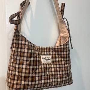 May include: A quilted hobo bag featuring a brown, black, and cream plaid pattern. The bag has a long shoulder strap with tie details at the top and a rectangular white fabric label in the center with the text SEW threaded printed in black.