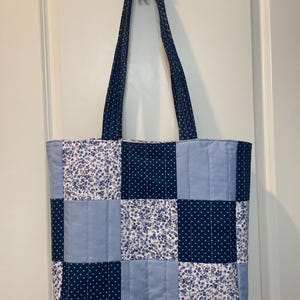May include: A quilted tote bag featuring a patchwork design of blue and white fabrics. The pattern alternates between solid light blue, dark blue with white dots, and a white fabric with a dense blue floral print. It has two long shoulder straps.