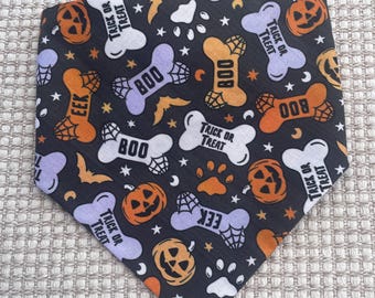 May include: A triangular pet bandana with a black background featuring a Halloween-themed pattern of orange jack-o'-lanterns, white and purple dog bones, paw prints, bats, and stars. The bones are printed with the text BOO, EEK, or TRICK OR TREAT.