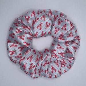 Op de afbeelding: Een grote, ronde scrunchie met een herhalend patroon van rode kreeften op een lichtblauwe en witte gestreepte achtergrond. De stof is gerimpeld en ziet er zacht uit, waardoor een volumineus haaraccessoire ontstaat.