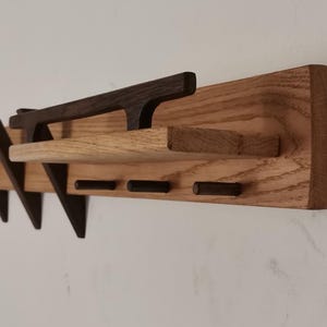 May include: A wooden wall-mounted coat rack with a shelf and multiple angled hooks. The rack is made of light brown wood with dark brown hooks and a shelf.