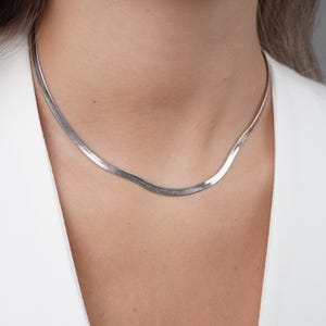 May include: A silver herringbone chain necklace. The necklace is flat and flexible, with a smooth, reflective surface. The chain is worn around the neck, resting on the chest. The background is a white blazer.