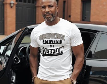 May include: White t-shirt with the text "OBEDIENCE ACTIVATED MY OVERFLOW" in a black and gray design. The person is wearing khaki pants and a gold watch. The background includes a black car and a brick building.