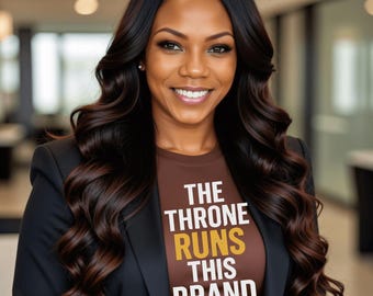 May include: A woman with long, dark wavy hair smiles, wearing a brown t-shirt with the text "THE THRONE RUNS THIS BRAND" in white and gold. She is also wearing a black blazer.
