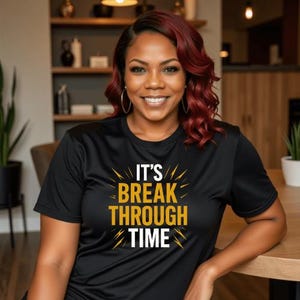 May include: Black t-shirt with the text "IT'S BREAK THROUGH TIME" in gold and white. The shirt is worn by a person with red hair and gold hoop earrings. The background includes a wooden table and shelves.