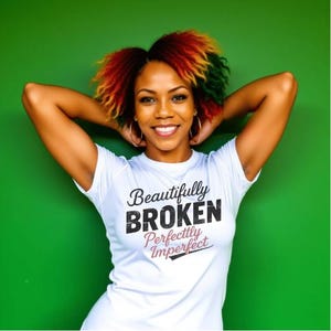 May include: A woman wearing a white t-shirt with the text "Beautifully BROKEN Perfectly Imperfect" in black and pink. Her hair is styled with red, yellow, and green highlights. She is smiling with her arms behind her head against a green background.