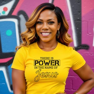May include: A woman wearing a yellow t-shirt with the text "THERE IS POWER IN THE NAME OF Jesus" in black. The woman is smiling, with gold hoop earrings and wavy hair. The background features a colorful graffiti wall.