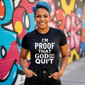 May include: Black t-shirt with the text "I'M PROOF THAT GOD DOESN'T QUIT" in white. The person is wearing black jeans and has blue hair. The background is a colourful graffiti wall.