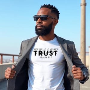 Puede incluir: Camiseta blanca con la palabra "TRUST" en negro y el texto "PSALM 91:2" debajo. La persona lleva una blazer gris y gafas de sol negras. El fondo es un cielo azul claro.