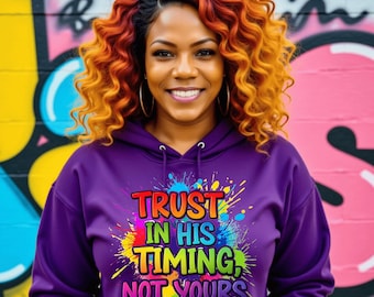 May include: A woman wearing a purple hoodie with the text "TRUST IN HIS TIMING, NOT YOURS" in a colorful, paint-splatter design. She has curly, orange and brown hair and is smiling. The background is a graffiti wall.
