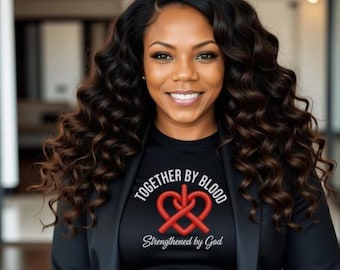 May include: A woman wearing a black blazer over a black t-shirt. The t-shirt has the words "TOGETHER BY BLOOD" and "Strengthened by God" printed on it, along with a red heart-shaped design. The woman has long, wavy dark hair.