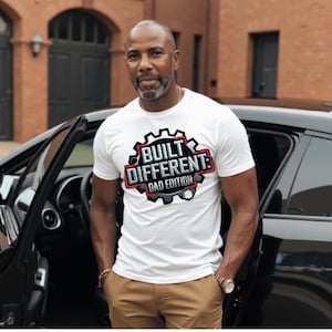 Pode incluir: Camiseta branca com o texto "BUILT DIFFERENT DAD EDITION" em um design preto, vermelho e cinza. A camiseta é de manga curta e é usada por uma pessoa ao lado de um carro preto.
