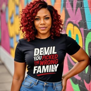Puede incluir: Una camiseta negra con el texto "DEVIL YOU PICKED THE WRONG FAMILY" en blanco y rojo. La persona lleva vaqueros azules y tiene el pelo rizado rojo. El fondo es una pared de graffiti colorida.