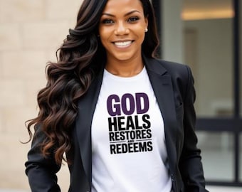 Può includere: Una donna indossa una maglietta bianca con la scritta "GOD HEALS RESTORES AND REDEEMS" in viola e nero. Indossa anche un blazer nero. La donna ha lunghi capelli scuri e ondulati e sta sorridendo.