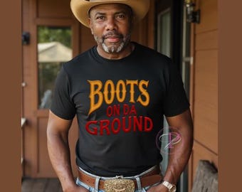 May include: A person wearing a black t-shirt with the text "BOOTS ON DA GROUND" in orange and red lettering. The person is wearing a brown cowboy hat and blue jeans with a belt buckle.