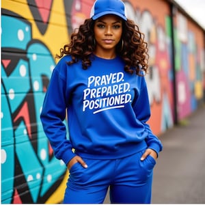 Puede incluir: Una mujer con un chándal azul y una gorra de béisbol azul con la parte trasera de malla blanca. La sudadera tiene la frase "PRAYED. PREPARED. POSITIONED." en blanco. El fondo presenta arte de graffiti colorido.