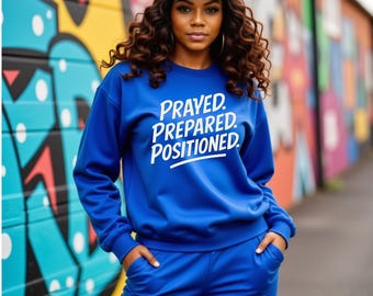 May include: A woman wearing a blue sweatsuit and a blue baseball cap with a white mesh back. The sweatshirt has the words "PRAYED. PREPARED. POSITIONED." in white. The background features colorful graffiti art.