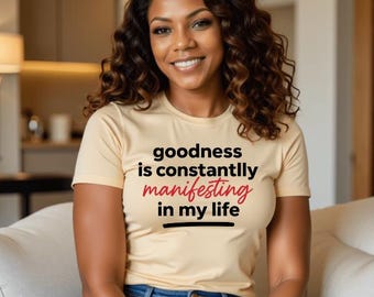 May include: A woman wearing a light beige t-shirt with the text "goodness is constantly manifesting in my life" printed in black and red. She is sitting on a white couch and wearing blue jeans.