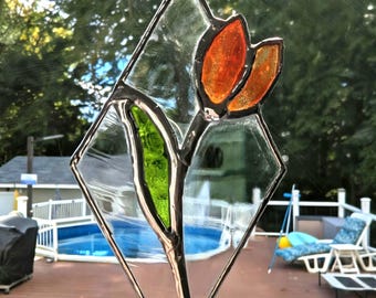 May include: A diamond-shaped stained glass suncatcher with a flower design. The flower has orange petals and a green leaf, set within a clear glass panel and a silver-coloured metal frame. It hangs from a chain. The background shows a garden with a swimming pool.