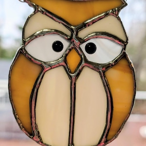 May include: Stained glass owl suncatcher with a chain for hanging. The owl features a design with yellow, cream, and white glass pieces. The eyes are black, and the glass is outlined with a dark metal. A decorative piece for windows.