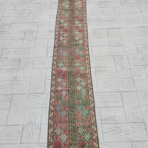 Könnte beinhalten: Ein langer, schmaler Teppich mit traditionellem türkischem Design. Der Teppich hat einen cremefarbenen Hintergrund mit einem sich wiederholenden Muster aus grünen, rosa und blauen Blumen. Der Teppich hat einen schwarzen Rand mit einem sich wiederholenden Muster aus kleinen, weißen Blumen.