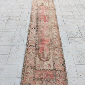 May include: A rectangular, vintage-style runner rug with a central red and pink design, framed by a border of brown and beige geometric patterns. The rug is placed on a light-colored, textured surface.