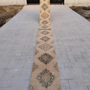 May include: A long, beige runner rug with a traditional Persian design. The rug has a green and brown border and features intricate floral patterns in the center.