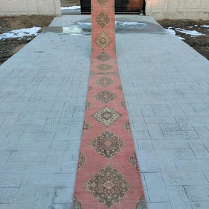 May include: A long, pink runner rug with a traditional Persian design. The rug has a repeating pattern of intricate floral motifs and geometric shapes in shades of brown, beige, and green.