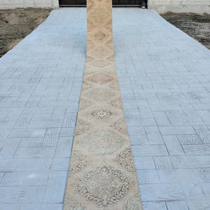 May include: A long, beige runner rug with a traditional floral pattern in shades of brown and green. The rug has a fringed edge and is laid out on a gray concrete patio.