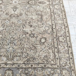 May include: A beige and brown area rug with an intricate floral pattern. The rug features a central design of stylized flowers and leaves, bordered by a detailed edge. The color palette is neutral, with shades of beige, brown, and gray.