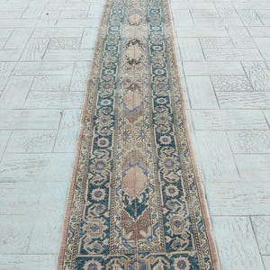 May include: A long, narrow rug with an intricate pattern. The rug features a central beige field with repeating geometric designs in shades of blue, brown, and red. The border is a darker blue with floral motifs. The rug is likely made of wool.