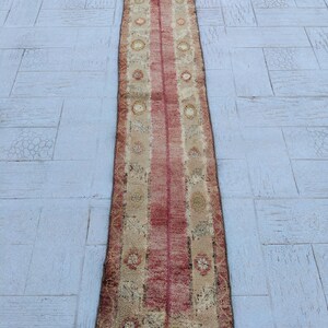 Könnte beinhalten: Ein langer, rechteckiger Teppich mit einem roten Mittelstreifen und beigen Rändern. Der Teppich weist ein sich wiederholendes Muster aus floralen und geometrischen Designs in Rot-, Beige- und Brauntönen auf. Der Teppich liegt auf einer hellgrauen Fliesenoberfläche.