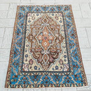 Puede incluir: Alfombra rectangular con un diseño central de medallón marrón, bordeada por un patrón azul claro y crema. La alfombra presenta motivos florales y geométricos en tonos marrón, naranja y azul, creando una estética vintage.