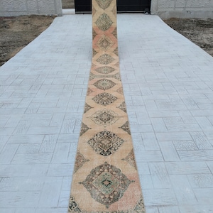 May include: A long, beige runner rug with a traditional Persian design. The rug has a repeating pattern of green, brown, and pink diamonds. The rug is laid out on a grey concrete surface.