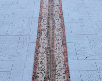 May include: A long, narrow rug with a cream background and a repeating pattern of pink, green, and black floral motifs. The rug has a fringe border.