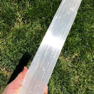 May include: A long, translucent white selenite crystal wand with a fibrous, striated texture. The crystal has a rectangular, columnar shape with a natural, slightly uneven finish along the edges and a reflective, pearlescent surface.