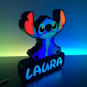 以下が含まれることがあります： 青と赤のスティッチのキャラクターのナイトライトで、ベースに「LAURA」という名前が書かれています。