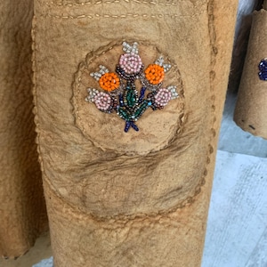 Puede incluir: Primer plano de una chaqueta de cuero marr&oacute;n con un bolsillo y abalorios florales. El dise&ntilde;o de cuentas presenta cuentas naranjas, rosas y verdes dispuestas en un patr&oacute;n de ramo de flores. La chaqueta tiene una superficie texturizada y costuras visibles.