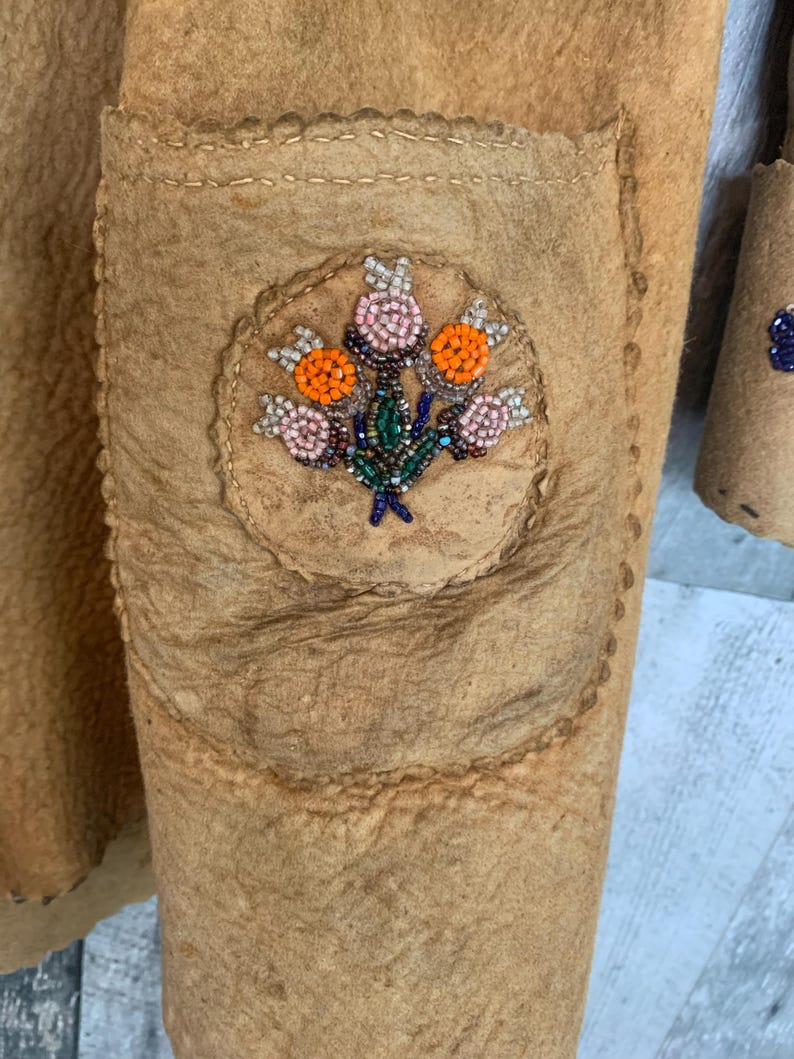 Puede incluir: Primer plano de una chaqueta de cuero marr&oacute;n con un bolsillo y abalorios florales. El dise&ntilde;o de cuentas presenta cuentas naranjas, rosas y verdes dispuestas en un patr&oacute;n de ramo de flores. La chaqueta tiene una superficie texturizada y costuras visibles.