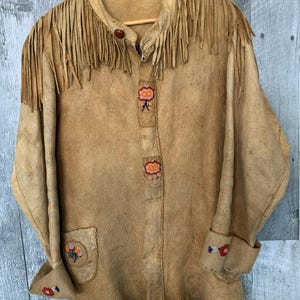 Puede incluir: Chaqueta de ante color canela con flecos en el cuello y los hombros. La chaqueta presenta bordados florales decorativos en los pu&ntilde;os, el bolsillo y la parte delantera. Colgada en una percha de madera.