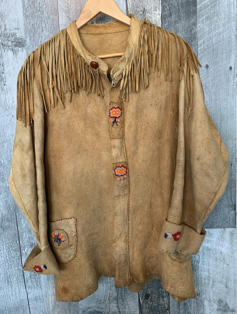 Puede incluir: Chaqueta de ante color canela con flecos en el cuello y los hombros. La chaqueta presenta bordados florales decorativos en los pu&ntilde;os, el bolsillo y la parte delantera. Colgada en una percha de madera.
