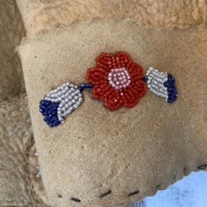 Puede incluir: Primer plano de un art&iacute;culo de ante color canela con un dise&ntilde;o floral de cuentas. La flor es roja con un centro rosa, y las hojas son blancas y azules. El art&iacute;culo tiene un borde cosido.